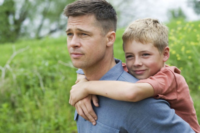 Medium shot of a clean-shaven Brad Pitt carrying a 13 year-old Laramie Eppler on his back in a green field